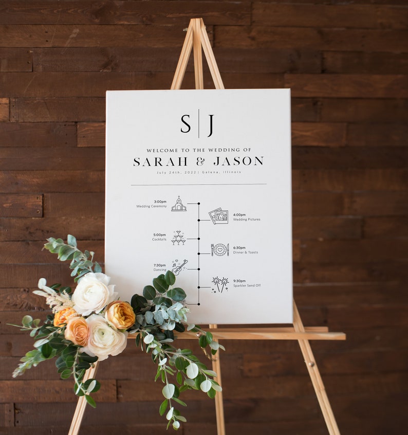 May include: A white wedding sign with a black easel stand. The sign has the initials "SJ" at the top and the text "Welcome to the wedding of Sarah & Jason" with the date "July 24th, 2022" and location "Galena, Illinois". The sign also has a timeline of wedding events with icons and times.