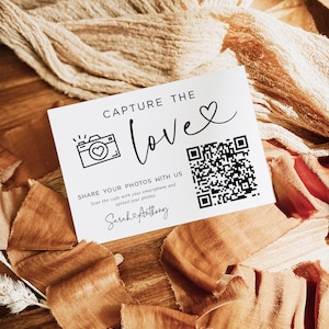 May include: White card with black text that reads "Capture the love" with a heart symbol. A camera icon with a heart inside is next to the text. A QR code is on the right side of the card. The text below the QR code reads "Share your photos with us. Scan the code with your smartphone and upload your photos. Sarah & Anthony"