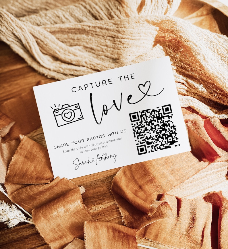 May include: White card with black text that reads "Capture the love" with a heart symbol. A camera icon with a heart inside is next to the text. A QR code is on the right side of the card. The text below the QR code reads "Share your photos with us. Scan the code with your smartphone and upload your photos. Sarah & Anthony"