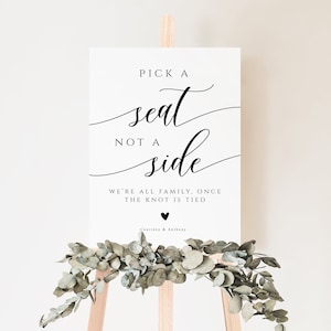 May include: White sign with black calligraphy that reads "Pick a seat, not a side. We're all family, once the knot is tied."  The sign is on a wooden easel with greenery.