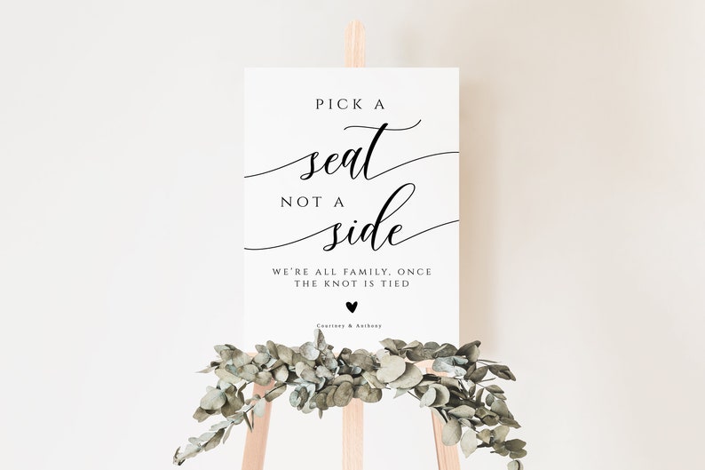 May include: White sign with black calligraphy that reads "Pick a seat, not a side. We're all family, once the knot is tied."  The sign is on a wooden easel with greenery.