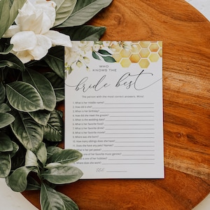 May include: A bridal shower game card with the text "Who Knows the Bride Best" in elegant script. The card features a honeycomb design and floral accents. The card is on a wooden surface, with greenery and a white flower in the background.