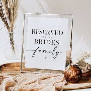 Puede incluir: Un letrero enmarcado con el texto "RESERVED FOR THE BRIDES family" en escritura negra. El letrero está en un marco de vidrio transparente, apoyado en una bandeja de madera con tela beige y jarrones decorativos.
