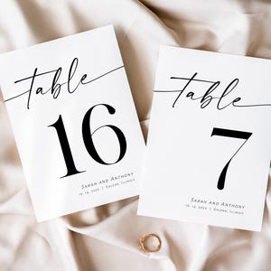 May include: Two white table number cards with black calligraphy. The top card says "Table 16" and the bottom card says "Table 7". Both cards have the text "Sarah and Anthony" and the date "10.16.2021" in smaller font.