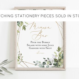 May include: A framed sign with the words "Mimosa Bar" in elegant gold script. The sign includes instructions: "Pour the Bubbly, Splash with some Juice, Garnish with Fruit." Greenery and gold geometric accents frame the text. The text "Matching Stationery Pieces Sold in Store" is at the top.