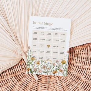 May include: A bridal bingo card with a floral design. The card has a grid with squares containing various wedding gift ideas, such as dish cloths, a cake stand, a laundry hamper, a table cloth, plates, mixing bowls, ramekins, baking pans, a gift card, drinking glasses, cash, a coffee pot, a heart, a sheet set, a whisk, napkins, measuring cups, a slow cooker, a toaster, a picture frame, wine glasses, a chopping board, coffee mugs, knives, and a vase.