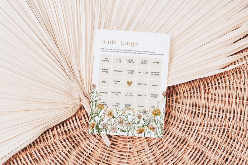 May include: A bridal bingo card with a floral design. The card has a grid with squares containing various wedding gift ideas, such as dish cloths, a cake stand, a laundry hamper, a table cloth, plates, mixing bowls, ramekins, baking pans, a gift card, drinking glasses, cash, a coffee pot, a heart, a sheet set, a whisk, napkins, measuring cups, a slow cooker, a toaster, a picture frame, wine glasses, a chopping board, coffee mugs, knives, and a vase.