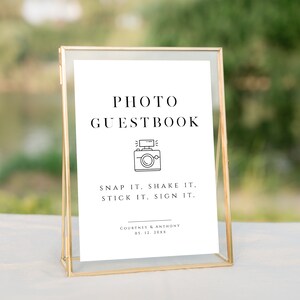May include: A gold-framed sign with white paper inside. The sign reads "PHOTO GUESTBOOK" with a camera icon and the text "SNAP IT, SHAKE IT, STICK IT, SIGN IT." Below the text is "COURTNEY & ANTHONY 01.12.208K".
