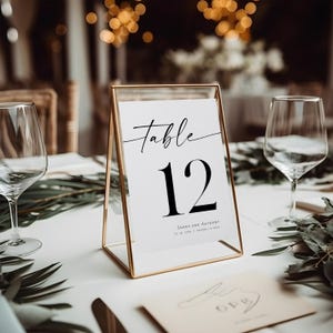 May include: A gold-framed table number card with the text "Table 12" in black font. The card also includes the names "Sarah and Anthony" and the date "10.14.2023" in smaller font.