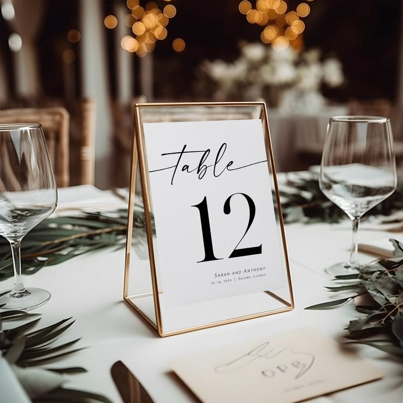 May include: A gold-framed table number card with the text "Table 12" in black font. The card also includes the names "Sarah and Anthony" and the date "10.14.2023" in smaller font.