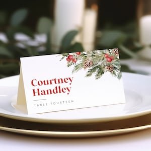 Puede incluir: Una tarjeta de lugar blanca con un diseño de acuarela de ramas de pino, piñas y bayas rojas. El texto en la tarjeta dice "Courtney Handley" y "Mesa Catorce".