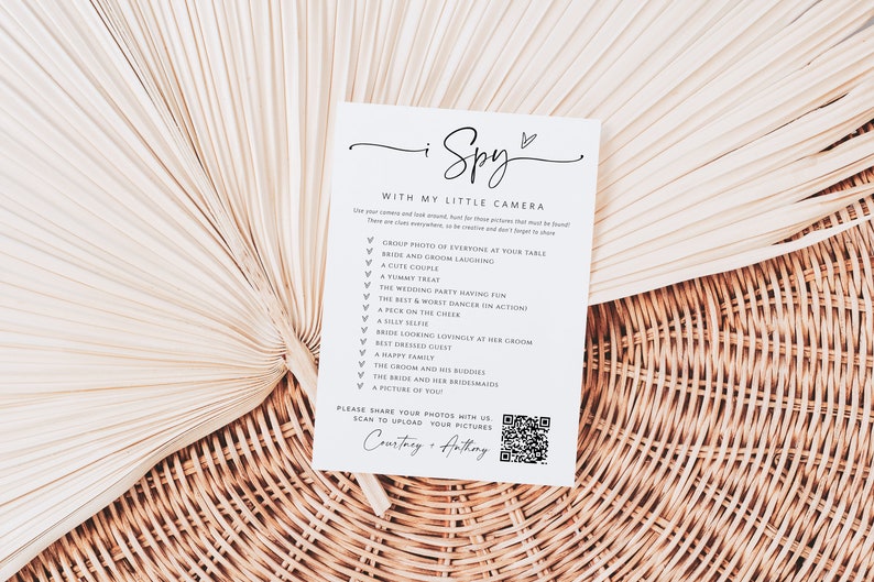 May include: A white card with the text "I Spy with my little camera" and a list of things to find at a wedding. The card also includes a QR code with the text "Please share your photos with us. Scan to upload your pictures. Courtney + Anthony."