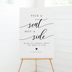 May include: White sign with black text that reads "Pick a seat, not a side. We're all family, once the knot is tied."  The sign also includes a small black heart and the names "Courtney & Anthony".