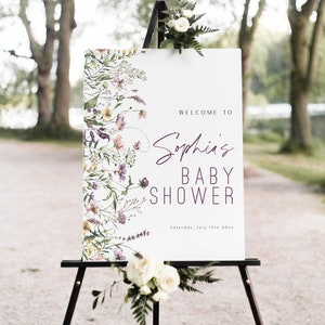 May include: Welcome to Sophia's Baby Shower sign with a floral design in shades of purple, pink, and yellow. The sign is on an easel and has the text "Welcome to Sophia's Baby Shower" and "Saturday, July 13th 20xx".