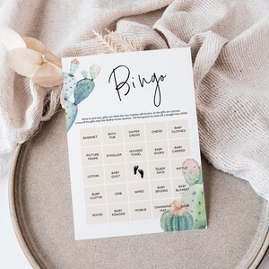 Puede incluir: Una tarjeta de bingo de baby shower imprimible con una ilustraci&oacute;n de cactus. La tarjeta tiene una cuadr&iacute;cula con casillas que contienen art&iacute;culos para beb&eacute;s como una cuna, una ba&ntilde;era, crema para pa&ntilde;ales, un body, ropa de beb&eacute;, un marco de fotos, un cochecito, una toalla con capucha, zapatos para beb&eacute;s, un portabeb&eacute;s, loci&oacute;n, una manta para beb&eacute;, un saco de dormir, un sonajero, pa&ntilde;ales, una cuna, toallitas h&uacute;medas, cucharas para beb&eacute;s, una manta para beb&eacute;, calcetines, talco para beb&eacute;s, un m&oacute;vil, una alfombra para cambiar pa&ntilde;ales y un libro.