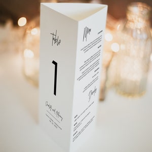 May include: White table number card with black text. The card is folded into a triangular shape. The number "1" is displayed in a large font. The text "Table" is written in cursive at the top of the card. The text "Charlie and Anthony" is written in cursive at the bottom of the card. The text "Toronto, Melbourne 08.21.2022" is written in a smaller font below the names.