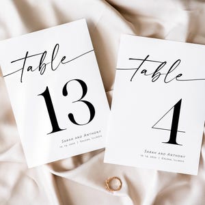 May include: Two white table number cards with black calligraphy. The top card says "Table 13" and the bottom card says "Table 4". Both cards have the names "Sarah and Anthony" and the date "10.14.2023" printed on them.