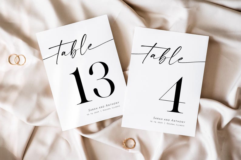 May include: Two white table number cards with black calligraphy. The top card says "Table 13" and the bottom card says "Table 4". Both cards have the names "Sarah and Anthony" and the date "10.14.2023" printed on them.