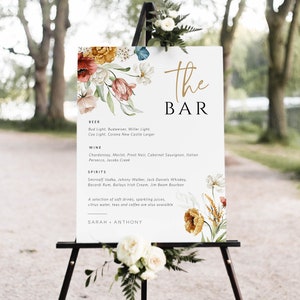 May include: A white sign with the words "The Bar" in gold lettering. The sign is decorated with watercolor flowers and lists the drinks available at the bar, including beer, wine, and spirits. The sign is on an easel with flowers on the bottom.