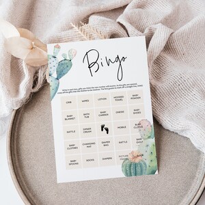 Puede incluir: Una tarjeta de bingo para baby shower imprimible con una ilustraci&oacute;n de cactus. La tarjeta tiene una cuadr&iacute;cula con casillas que incluyen art&iacute;culos que se suelen regalar en las baby showers, como pa&ntilde;ales, toallitas y una toalla con capucha. El texto "Bingo" est&aacute; en la parte superior de la tarjeta.