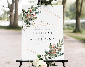 May include: Welcome sign with a gold geometric frame and greenery with red berries. The sign reads "Welcome to our wedding Hannah and Anthony 08.12.202x Galena | Illinois".