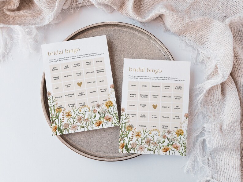 May include: Two bridal bingo cards with a floral design. The cards have a grid of squares with different wedding-related gifts listed in each square. The centre square of each card has a gold heart.
