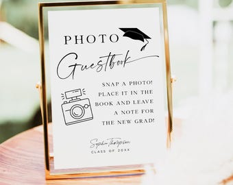 May include: A white sign with black text and a graduation cap graphic. The sign reads "PHOTO Guestbook SNAP A PHOTO! PLACE IT IN THE BOOK AND LEAVE A NOTE FOR THE NEW GRAD! Sophia Thompson CLASS OF 20XX"