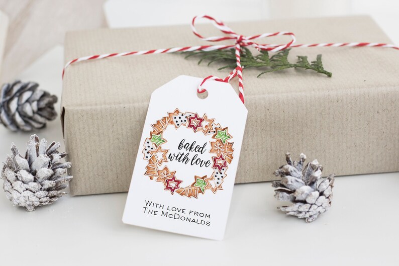 May include: White gift tag with a red and white twine bow. The tag has a hand-drawn illustration of a wreath made of gingerbread cookies with the text "baked with love". The text "With love from The McDonalds" is printed below the illustration.