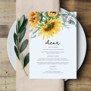 May include: A wedding menu card with a watercolor illustration of sunflowers and eucalyptus leaves. The menu includes a first course of goat cheese, red onion, and aubergine tart, a second course of filet mignon with sauteed mushrooms, roasted potatoes, and asparagus, and desserts of wedding cake and vanilla white chocolate mousse.
