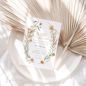 May include: A baby shower invitation featuring a floral wreath design. The text reads "Baby in Bloom" and includes the name "Courtney Laura" and the date "Sat 23 May".
