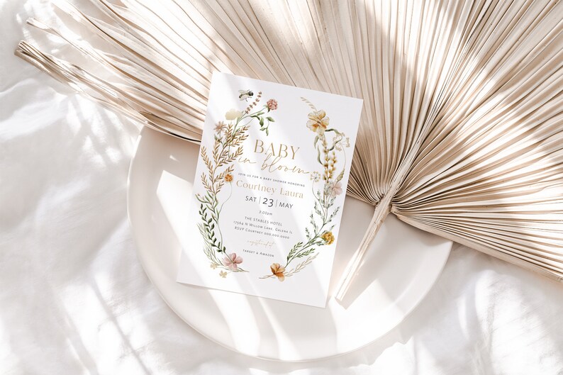 May include: A baby shower invitation featuring a floral wreath design. The text reads "Baby in Bloom" and includes the name "Courtney Laura" and the date "Sat 23 May".