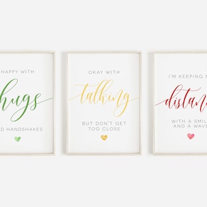 May include: Three framed prints with white backgrounds and light wood frames. The first print says "Happy with hugs and handshakes" in green text with a green heart. The second print says "Okay with talking but don't get too close" in yellow text with a yellow heart. The third print says "I'm keeping my distance with a smile and a wave" in red text with a pink heart.