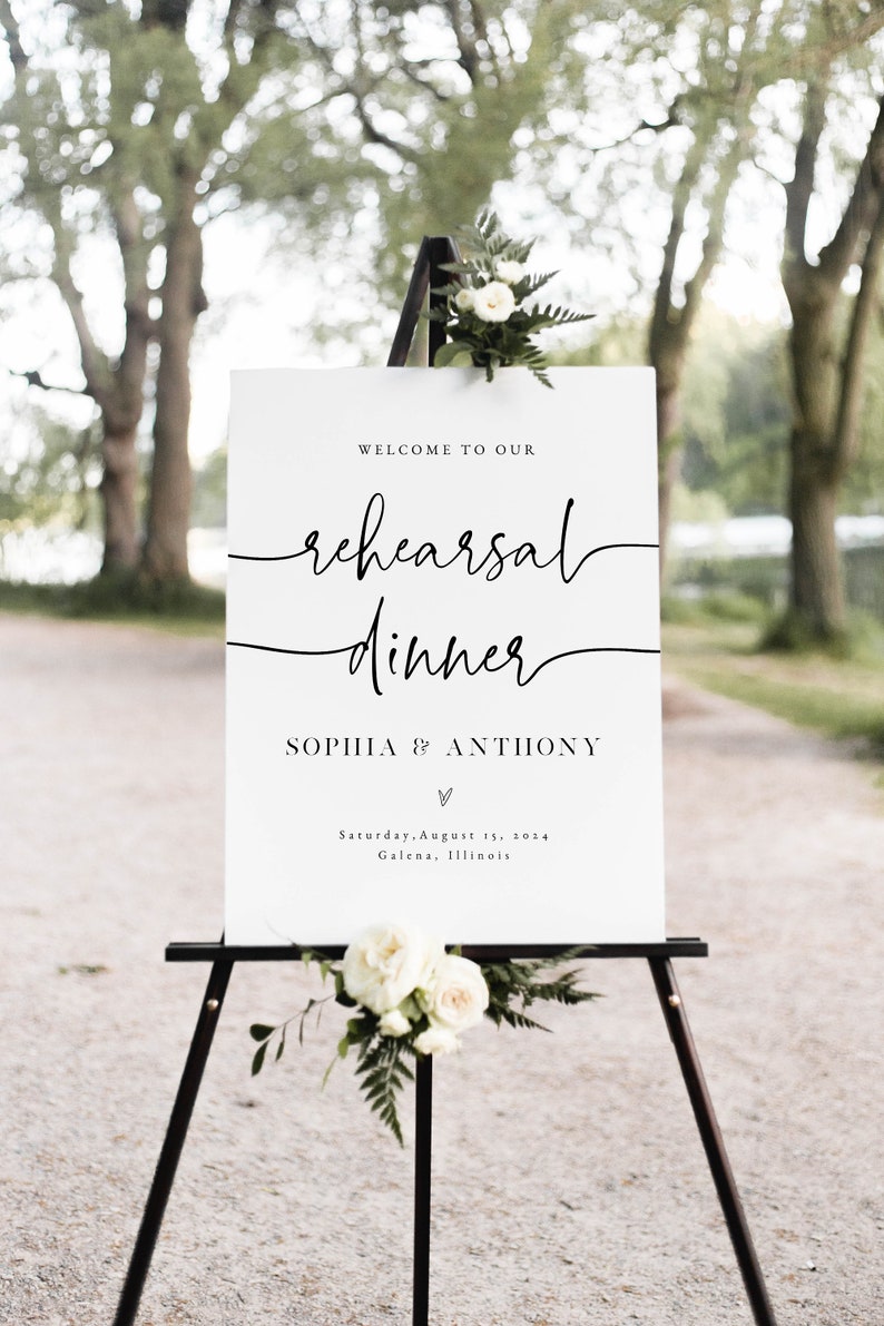 May include: White sign with black text on an easel. The sign reads "Welcome to our rehearsal dinner Sophia & Anthony" with a heart symbol and the date "Saturday, August 15, 2014" and location "Galena, Illinois".