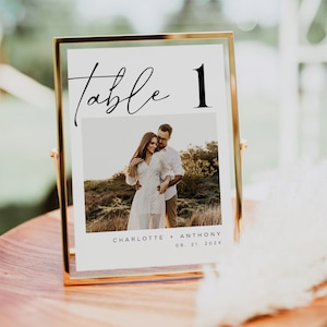 May include: A gold-framed table number sign with a white background. The number "1" is in black text and the word "table" is in black cursive script. A photo of a couple is below the table number. The text below the photo reads "Charlotte + Anthony 08.21.202X".