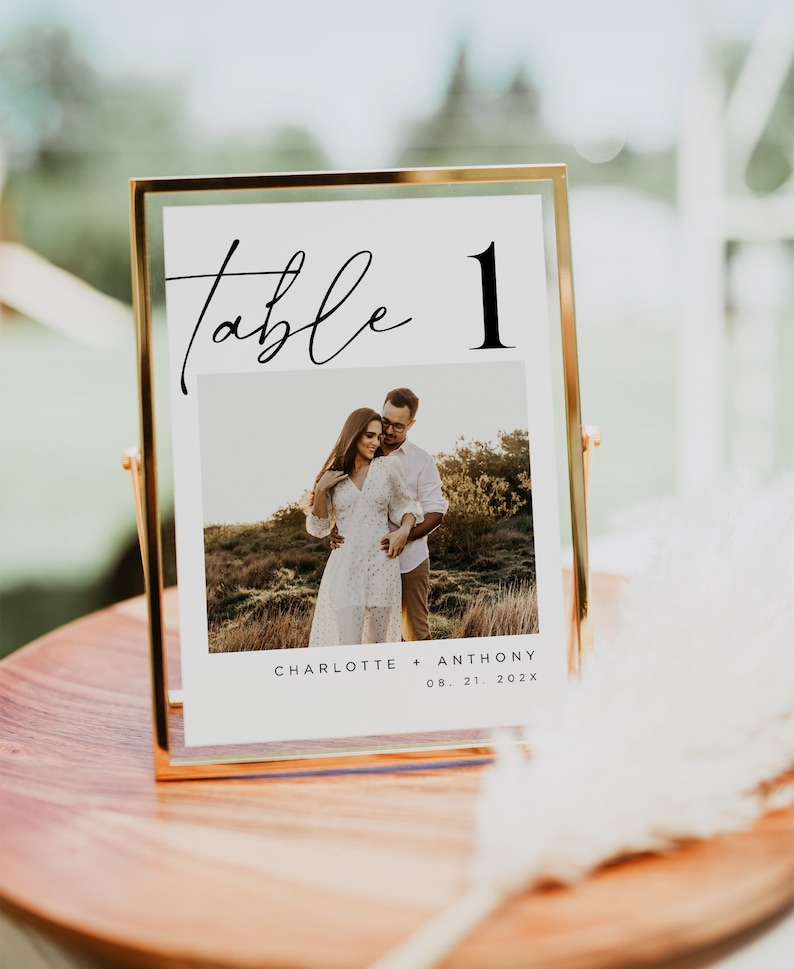 May include: A gold-framed table number sign with a white background. The number "1" is in black text and the word "table" is in black cursive script. A photo of a couple is below the table number. The text below the photo reads "Charlotte + Anthony 08.21.202X".
