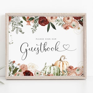 May include: A white sign with a floral border in shades of red, orange, and green. The sign says "Please sign our Guestbook" with a heart symbol. There are two pumpkins at the bottom of the sign.
