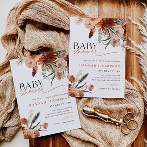 May include: Two baby shower invitations with a floral design in shades of orange, pink, and green. The invitations are on a wooden surface with a beige fabric overlay. The text on the invitations reads "Baby Shower" and includes the name "Hannah Thompson", the date "Sat | Nov 17 | 2PM", the location "The Smith Residence", and the address "276 Hillow Lane, Galena, IL".