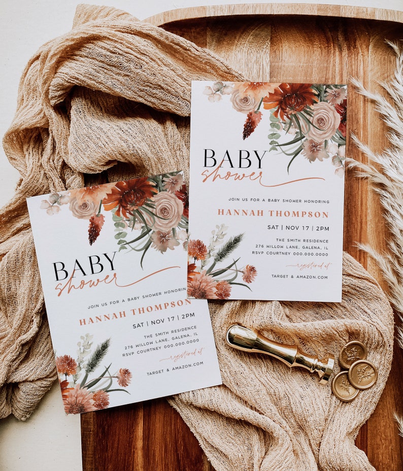 May include: Two baby shower invitations with a floral design in shades of orange, pink, and green. The invitations are on a wooden surface with a beige fabric overlay. The text on the invitations reads "Baby Shower" and includes the name "Hannah Thompson", the date "Sat | Nov 17 | 2PM", the location "The Smith Residence", and the address "276 Hillow Lane, Galena, IL".