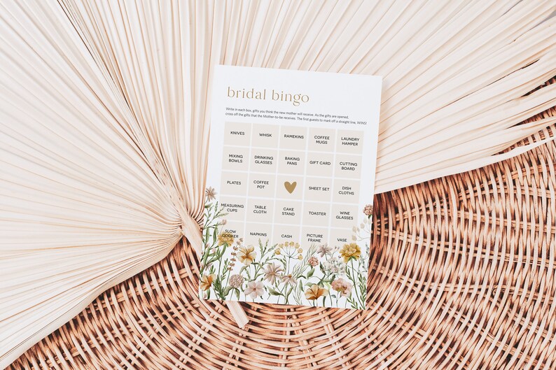 May include: A bridal bingo card with a floral design. The card has a grid of squares with various wedding gift ideas written in each square, such as knives, whisk, coffee mugs, and picture frame.