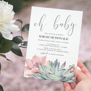 May include: Baby shower invitation with the words "oh baby" in elegant script. The invitation includes details for Sarah McDonald's baby shower, with a watercolor succulent design at the bottom. The invitation is held by a person.