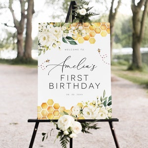 May include: Welcome to Amelia's First Birthday sign featuring a honeycomb pattern, white flowers, and bees. The date is 08.30.20XX.