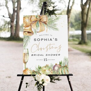 May include: A white sign with a gold and green watercolor design of Christmas ornaments, bells, and a bow. The text on the sign reads "Welcome to Sophia's Christmas Bridal Shower, Saturday 15 December".