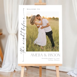 Può includere: Un cartello di benvenuto bianco su un cavalletto di legno. Il cartello presenta una foto di una coppia che si abbraccia in un campo. Il testo recita "Welcome to The night before" e i nomi "Amella & Landon" con la data e il luogo sottostanti.
