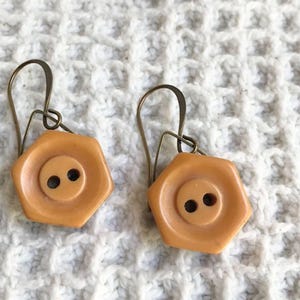 May include: A pair of unique earrings featuring light orange, hexagonal button-shaped pendants with two black holes in the center. The earrings have bronze-colored ear wires and are set against a white, textured background.