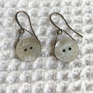 May include: A pair of earrings featuring white button-shaped charms with a crosshatch pattern and two black holes. The earrings are suspended from a brass wire.