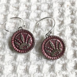 May include: A pair of dangle earrings with a circular, reddish-purple pendant. Each pendant features a silver-colored design of a crown or sunburst. The earrings have silver-colored ear wires and are displayed on a white, textured surface.