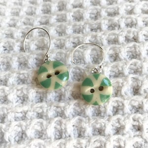May include: A pair of silver earrings with green and white ceramic button-shaped charms. The buttons have a swirl design and two holes.