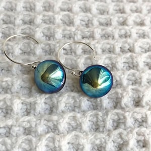 May include: A pair of iridescent blue and green orb earrings with silver-colored hooks. The earrings have a reflective, glossy finish, and are set against a white, textured background.