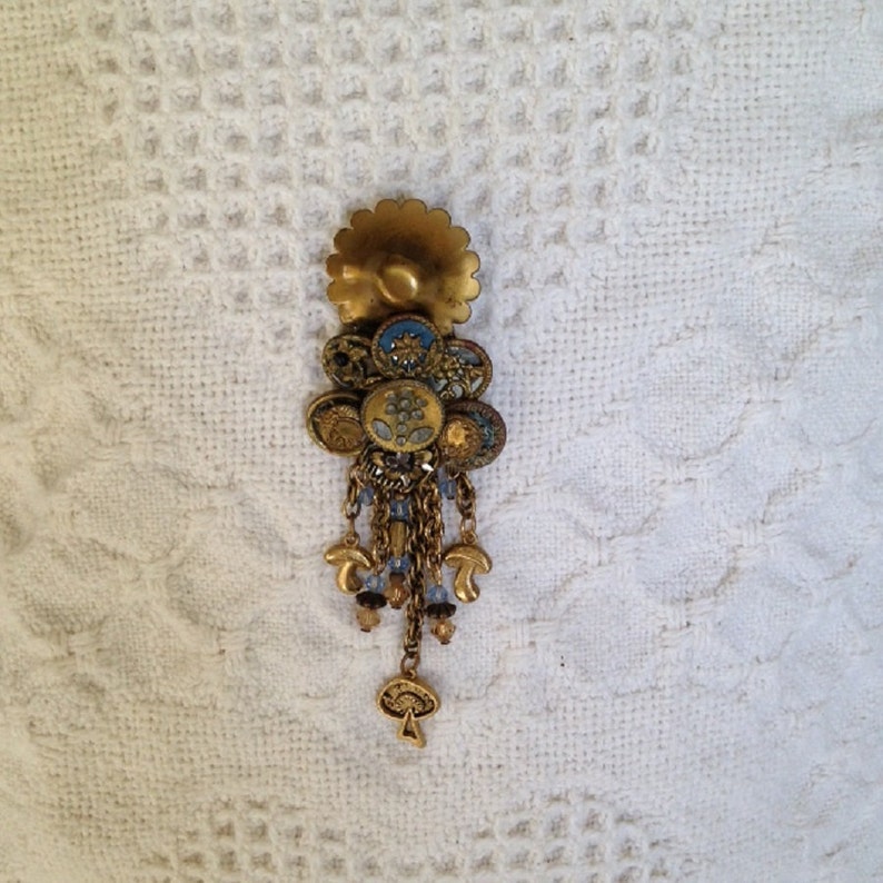 May include: A gold-toned flower-shaped brooch with blue and gold accents. The brooch has a dangling chain with blue beads and gold mushroom charms.