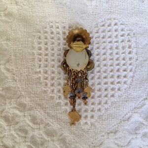 May include: A gold-toned brooch with a large, round, iridescent center surrounded by smaller, colorful stones. The brooch has a dangling chain with small, gold-toned charms.